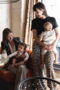Maman qui allaite debout adossée à une fenêtre qui sourit à une maman qui sourit à son enfant assise au restaurant Ganache à Bordeaux
