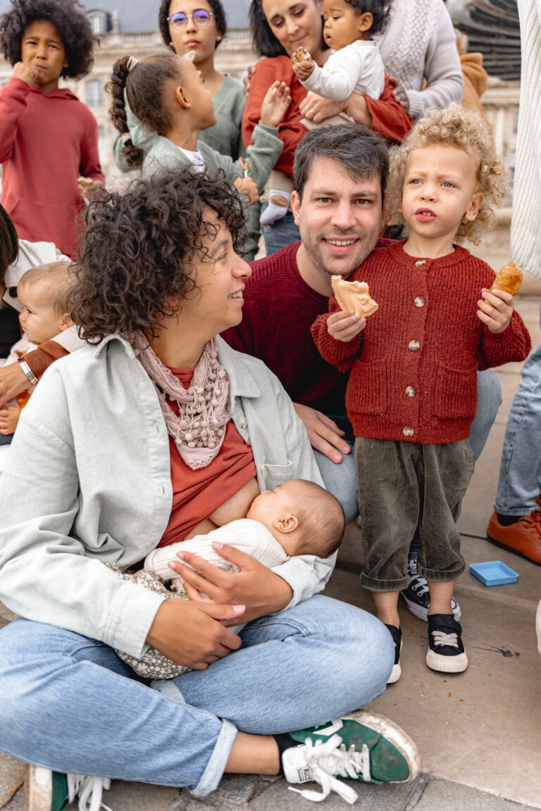 Maman assise en tailleur qui allaite son bébé en regardant son mari et son fils qui mangent des gâteaux lors de #jaifaimjemange 2024 à Bordeaux