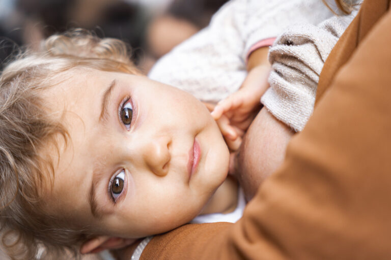 Petite fille qui sourit à l'objectif en tripotant le téton de sa mère qui l'allaite lors de #jaifaimjemange 2024 à Bordeaux