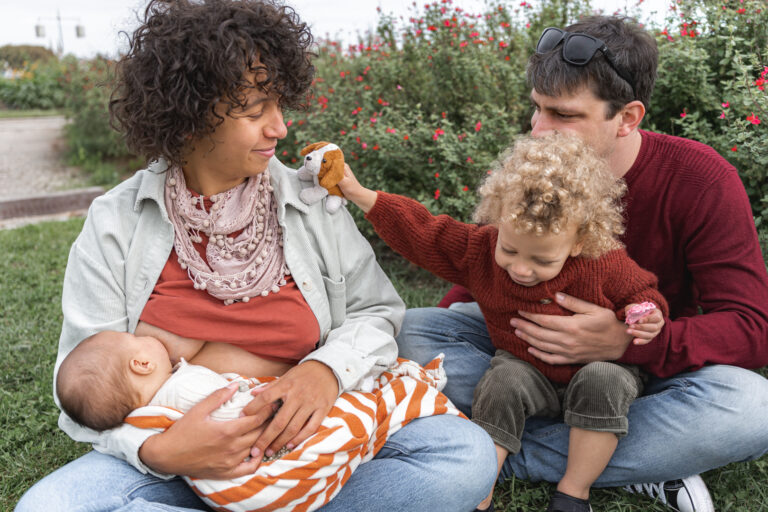 Maman qui allaite son bébé assise en tailleur dans l'herbe pendant que son mari tient leur fils qui montre son doudou lors de #jaifaimjemange 2024 à Bordeaux