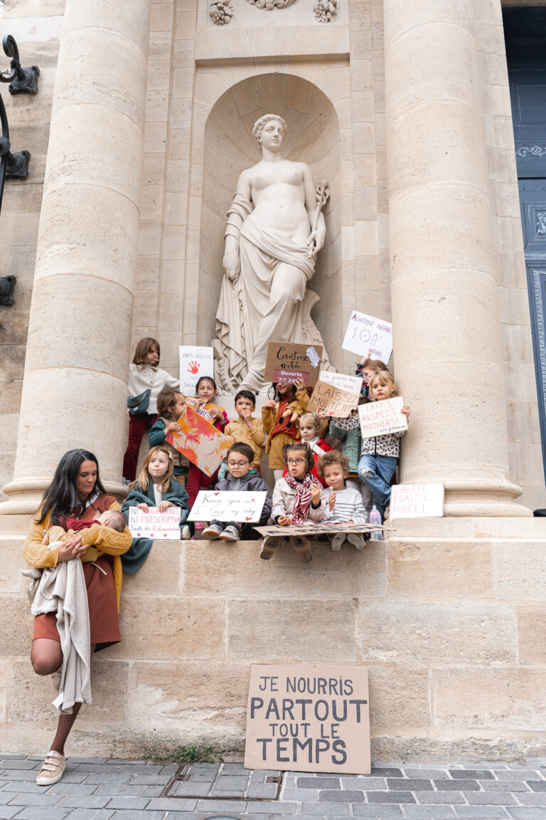 Maman qui allaite son bébé adossée contre une colonne de la mairie à côté d'une bande d'enfants qui tiennent des pancartes lors de #jaifaimjemange 2025 à bordeaux