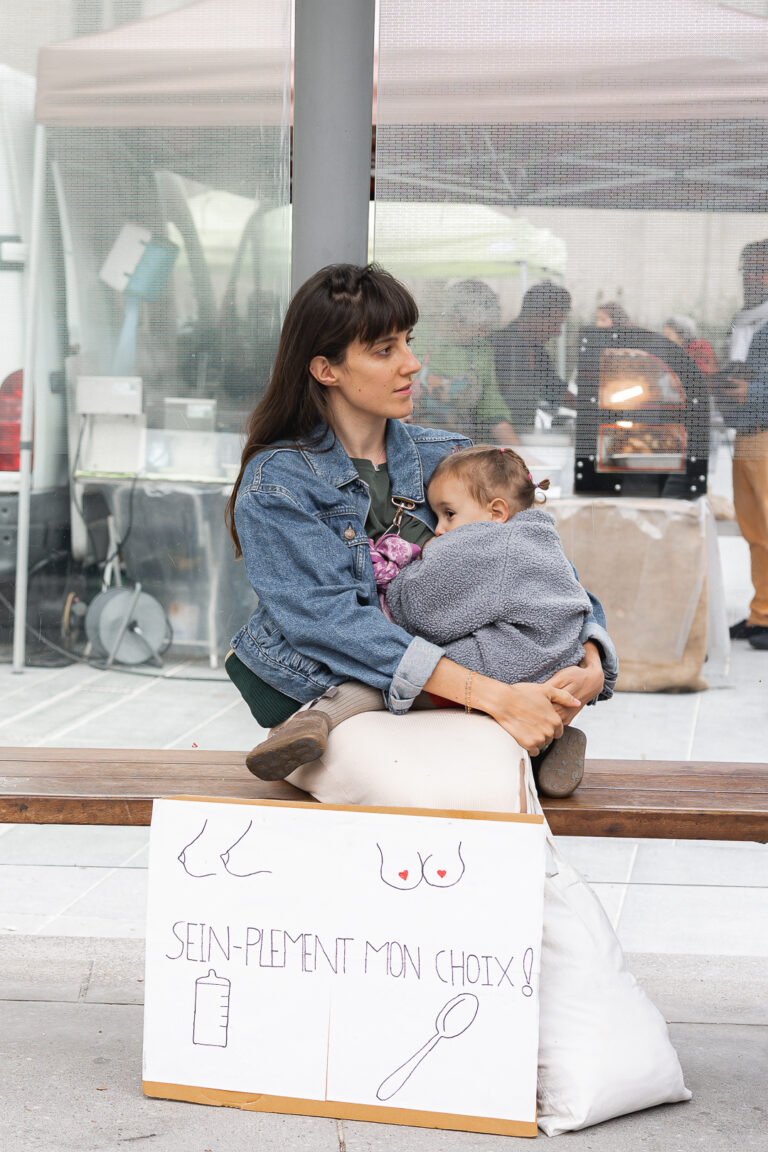 Maman qui allaite sa fille à un arrêt de tram, avec une pancarte pour l'allaitement maternel posée à ses pieds lors de #jaifaimjemange 2025 à Bordeaux