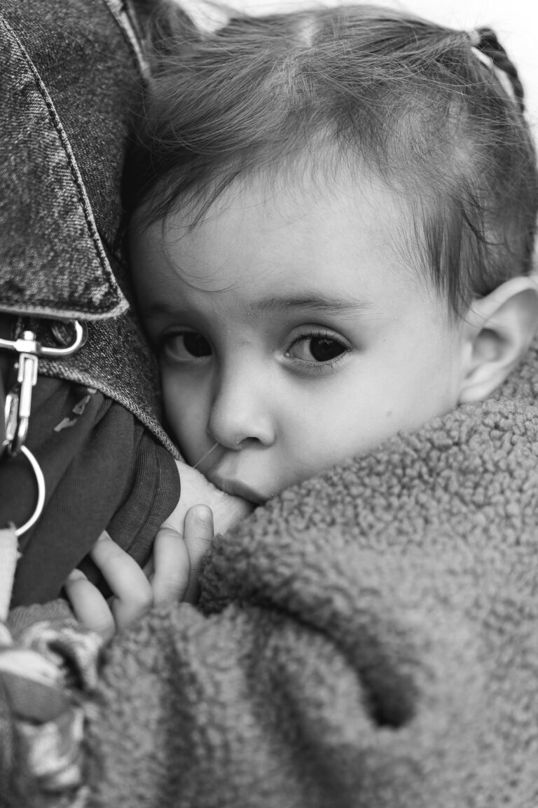 Photo en noir et blanc d'une petite fille qui regarde l'objectif en tétant le sein de sa mère dans le tram lors de #jaifaimjemange 2025 à Bordeaux