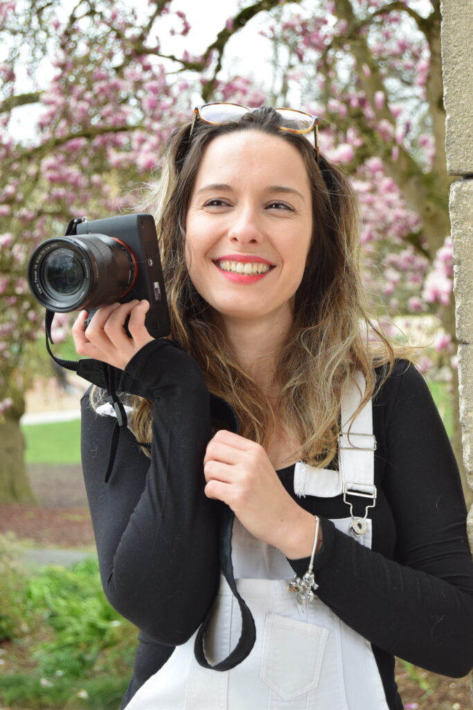 Photographe qui tient son appareil photo avec un grand sourire, lunettes sur la tête avec des cerisiers en fleurs en fond pour sa présentation