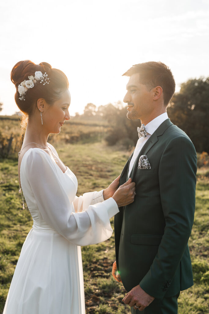 Jeune mariée rhabillant son époux lors de la séance couple de leur mariage dans les vignes au coucher du soleil