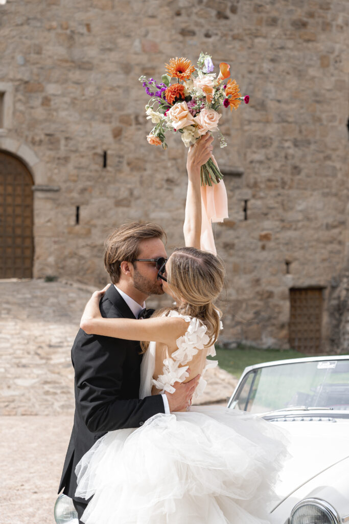 Jeunes mariés s'embrassant devant le château de Dio avec leur voiture de collection en levant le bouquet de la mariée en l'air