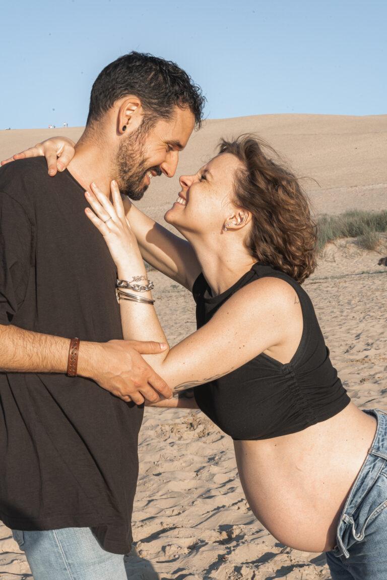 Homme qui regarde yeux dans les yeux sa femme enceinte suspendue à son cou en souriant lors d'une séance grossesse à la plage des dunes du pilat