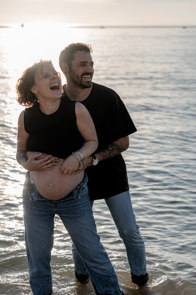 homme riant qui tient sa femme enceinte par derrière, morte de rire lors d'une séance grossesse à la plage des dunes du pilat au coucher du soleil