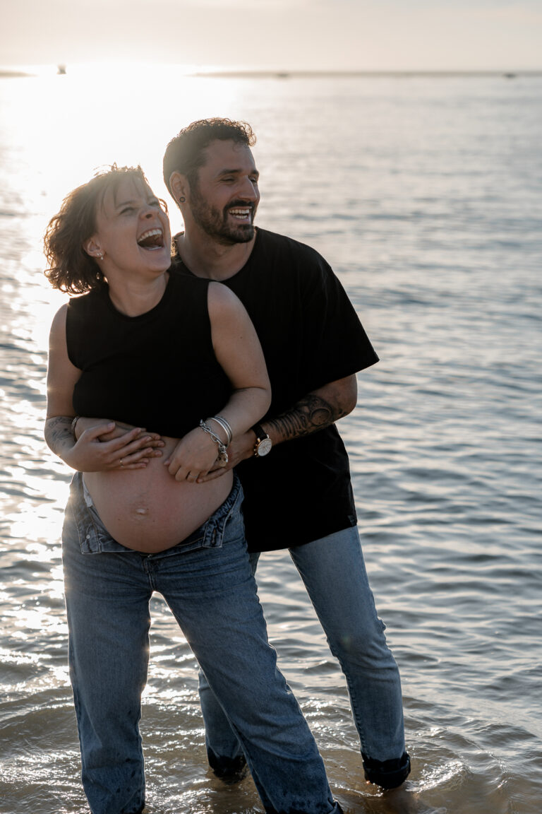 homme riant qui tient sa femme enceinte par derrière, morte de rire lors d'une séance grossesse à la plage des dunes du pilat au coucher du soleil