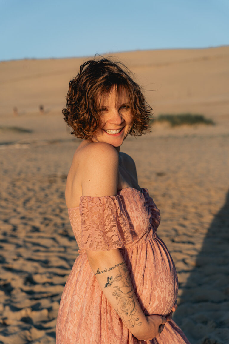 Femme enceinte qui rit face à l'objectif dans une robe en tenant son ventre lors d'une séance grossesse à la plage des dunes du pilat au coucher du soleil