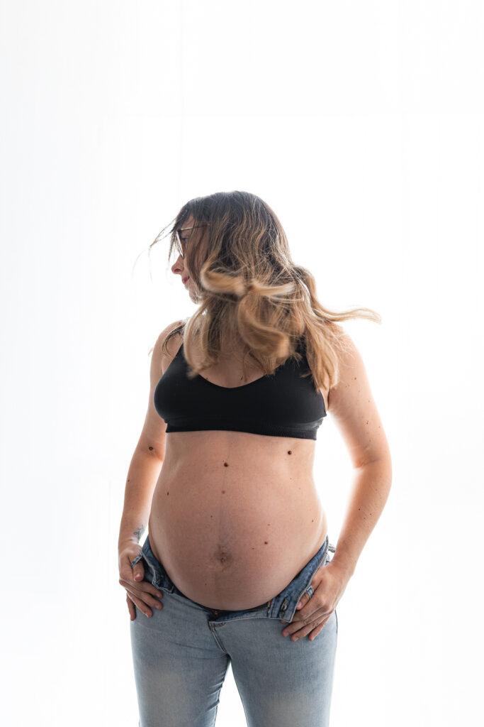Femme enceinte en brassière et jean qui fait voler ses cheveux pendant une séance grossesse au home studio à bordeaux
