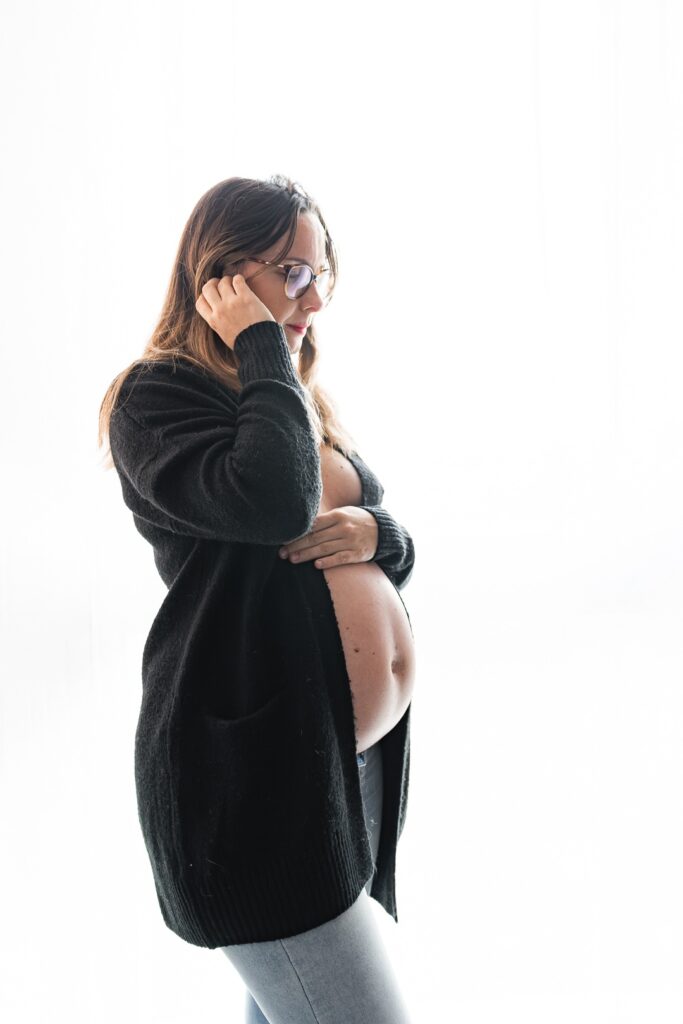 Femme enceinte songeuse en long gilet, ventre nu lors d'une séance grossesse au home studio à Bordeaux