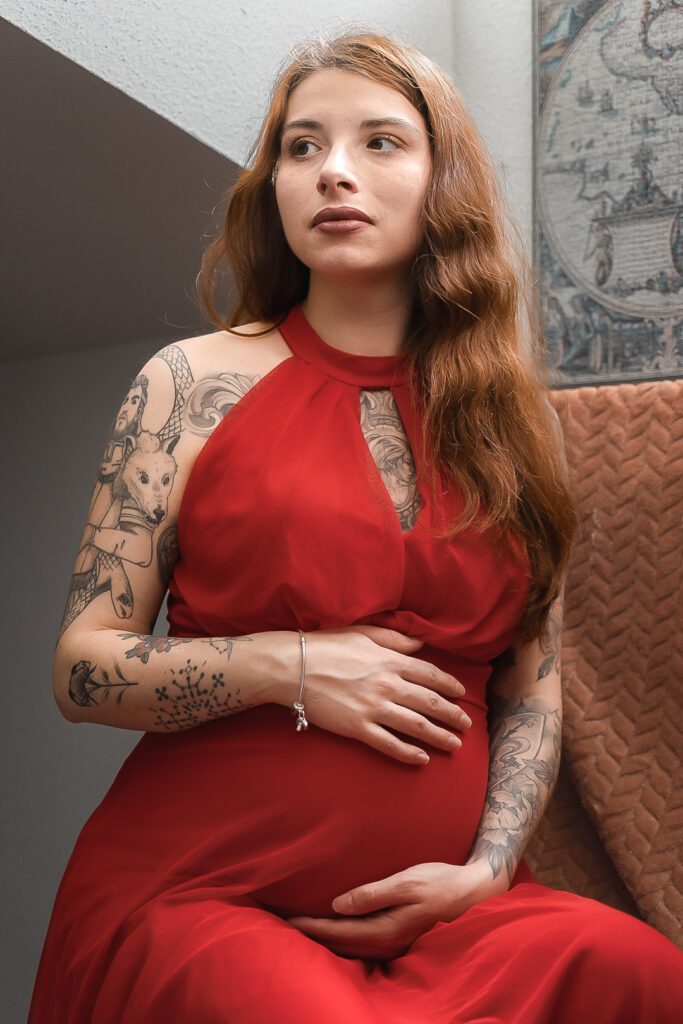 Femme enceinte dans une robe rouge touchant son ventre, le regard rêveur lors d'une séance grossesse à domicile à Blanquefort