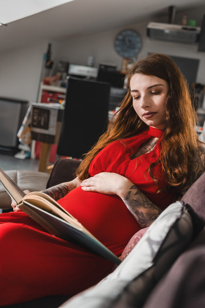 Femme enceinte qui lit un livre, assise sur son canapé, la main posée sur son ventre lors d'une séance grossesse à domicile à Blanquefort