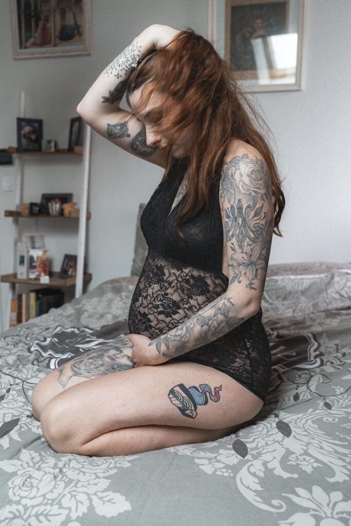 Femme enceinte en body qui se recoiffe assise sur son lit, la main sur son ventre lors d'une séance grossesse à domicile à Blanquefort
