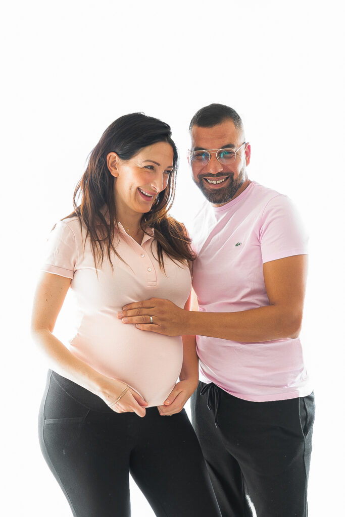 Femme qui regarde en dehors de l'objectif en riant, homme qui rit en regardant l'objectif en touchant son ventre lors d'une séance grossesse au Home Studio à Bordeaux