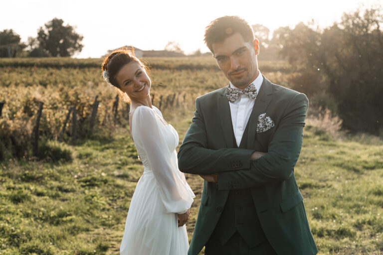 Marié bras croisé qui regarde l'objectif pendant que sa femme se moque gentiment derrière en robe de mariée lors de la séance photo couple de leur reportage de mariage au domaine au coucher du soleil dans les vignes