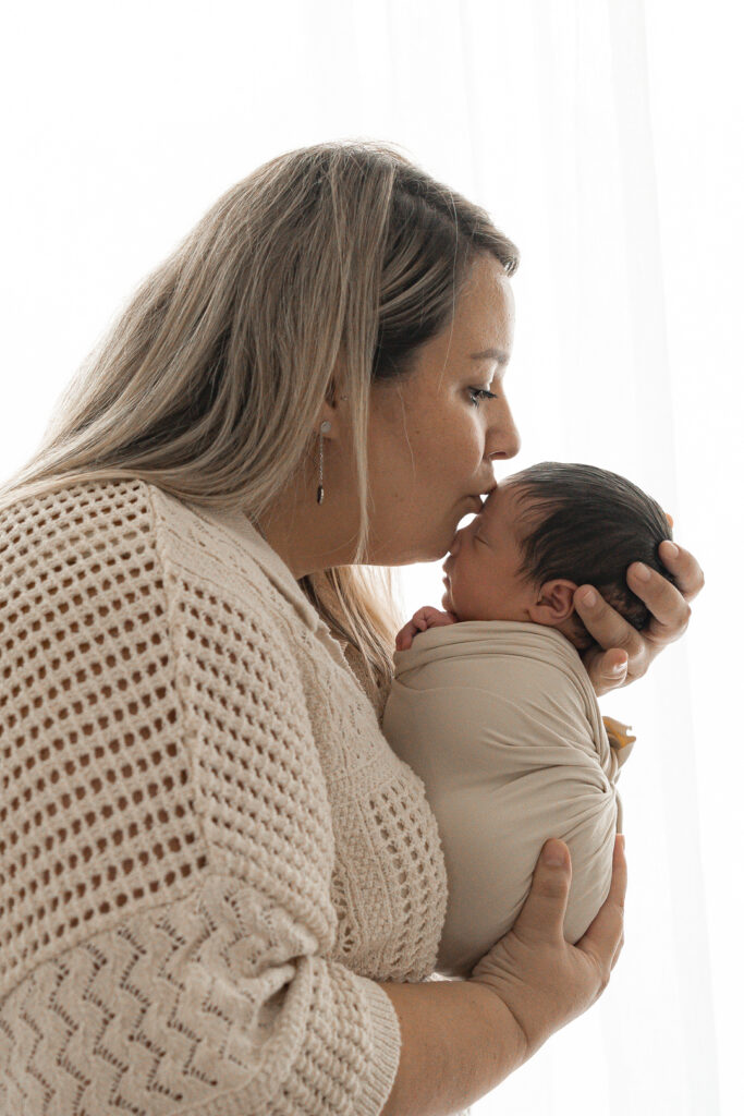 Bébé nouveau-né emmailloté dans les bras de sa maman qui l'embrasse sur le front lors d'une séance naissance à mon home studio sur Bordeaux