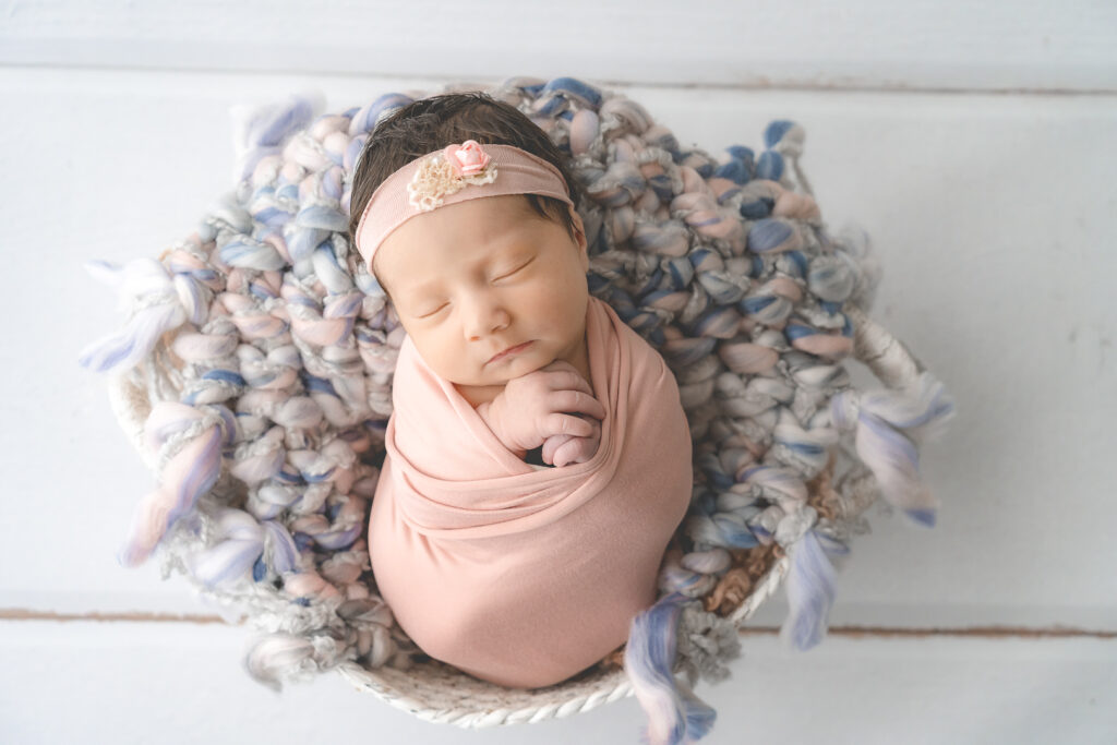 Bébé nouveau-né emmailloté dans un tissu rose et posé dans un panier lors d'une séance naissance en baby-posing à mon home studio sur Bordeaux