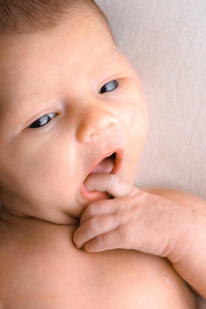 Gros plan sur un Bébé nouveau-né réveillé qui met son doigt dans la bouche pendant une séance naissance à mon home studio à Bordeaux