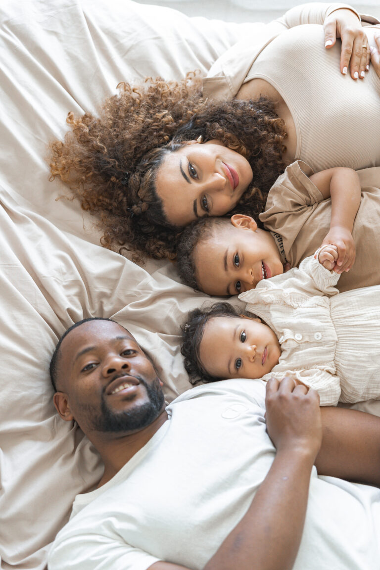 Famille allongée sur un lit et qui regarde la photographe en souriant lors d'une séance photo en famille à domicile à Pessac