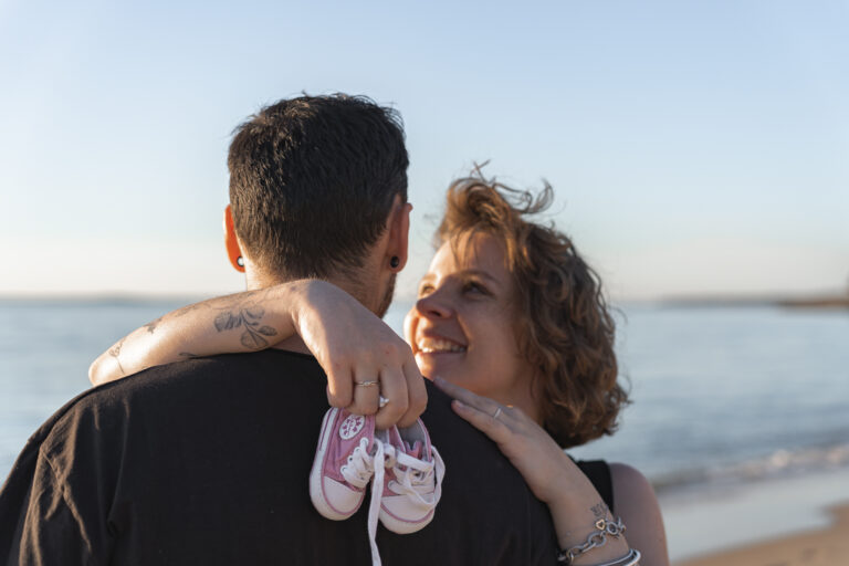 Future maman qui tient les premières basket de bébé en souriant au futur papa lors d'une séance photo grossesse en extérieur aux dunes du pilat sur le bassin d'Arcachon
