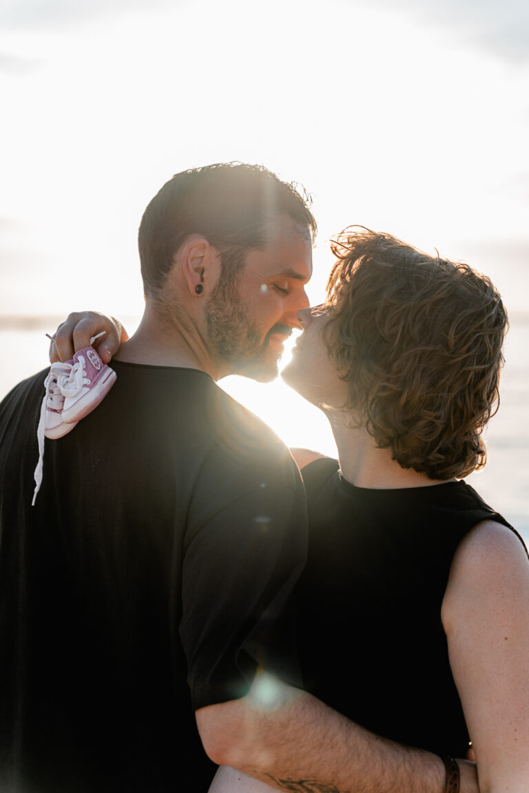 Futurs parents nez à nez, qui tiennent les premières basket de bébé lors de leur séance grossesse au coucher du soleil à la plage des dunes du pilat