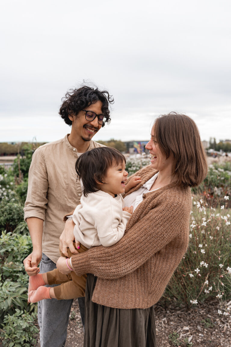 Maman qui tient son fils dans les bras en riant, papa les regarde en souriant lors d'une séance famille sur les quais de Bordeaux