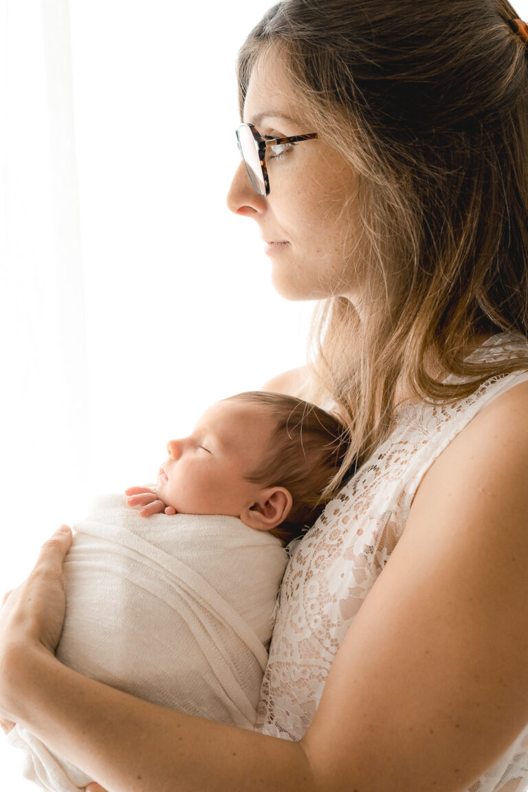 Bébé emmailloté dans un tissu blanc dans les bras de sa maman qui regarde au-dehors lors d'une séance nouveau-né en home studio à bordeaux