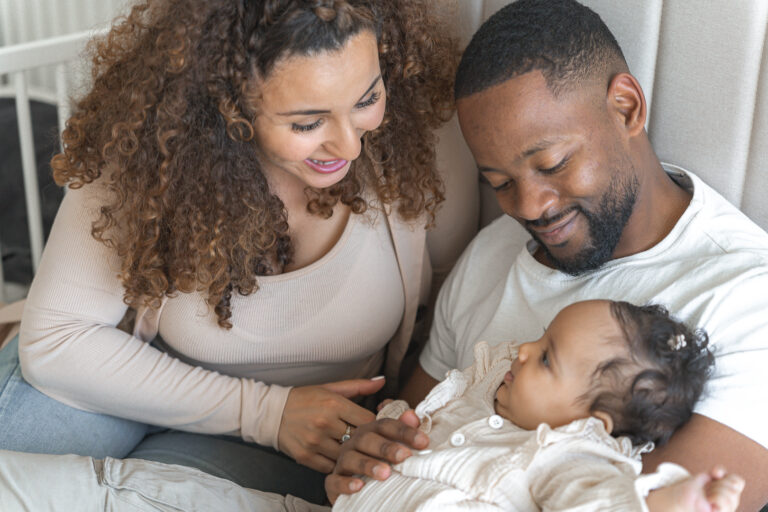 Bébé dans les bras de son papa pendant que maman le regarde en souriant durant un shooting naissance à domicile sur Pessac