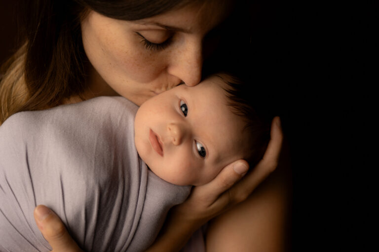 bébé réveillé et emmailloté dans les bras de sa maman qui lui fait un bisous sur la tempe lors d'un shooting naissance à mon home studio sur bordeaux