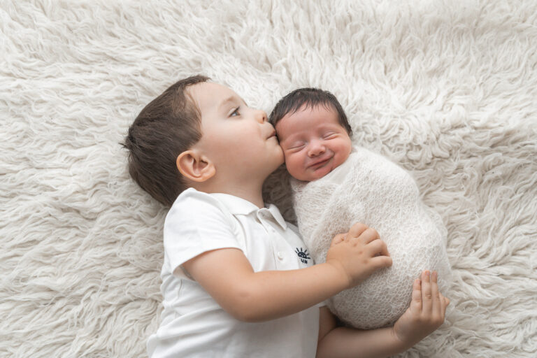 Bébé emmailloté entre les mains de son frère qui lui fait un bisou sur la tempe lors d'un shooting naissance à mon home studio sur bordeaux