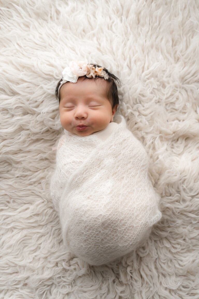 Nouveau-né emmailloté dans un tissu blanc avec un bandeau fleuri qui fait des bulles, posé sur un plaid lors d'un shooting bébé en home studio sur bordeaux
