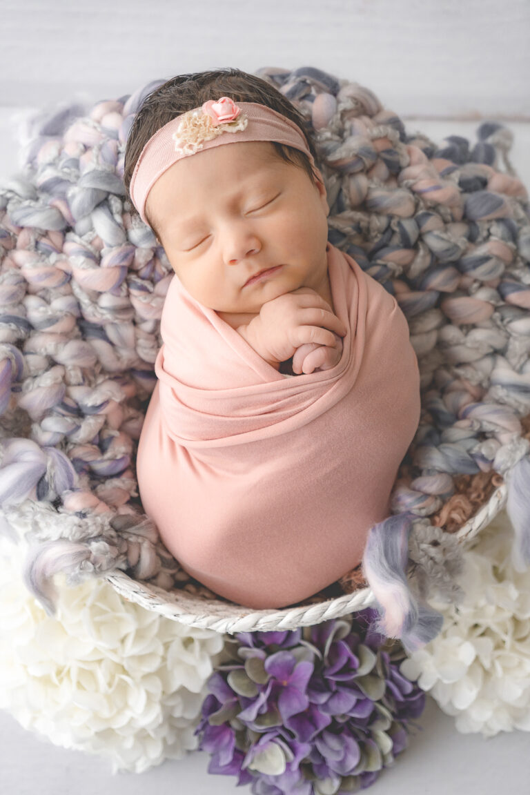mise en scène d'un nouveau-né emmailloté dans un tissu rose et posé dans un panier entouré de fleurs lors d'une séance photo de naissance en home studio sur bordeaux