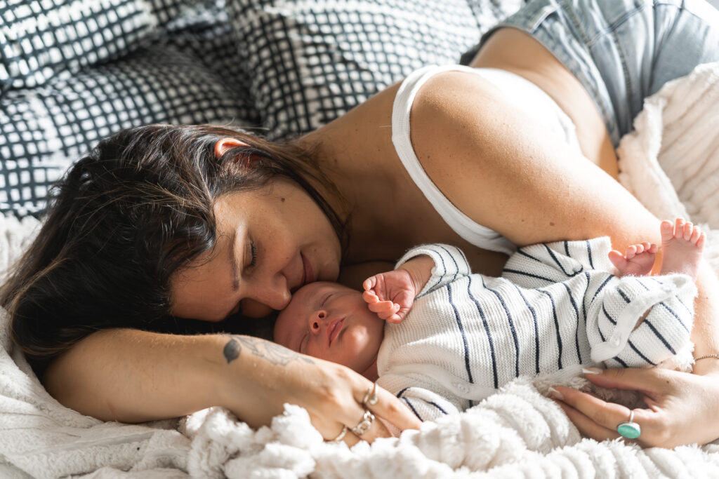 Maman qui sourit allongée dans un lit, la tête posée sur celle de son nouveau-né lors d'une séance naissance à domicile à Mios sur le bassin d'arcachon