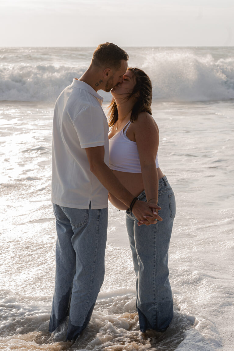Futurs parents qui s'embrassent les pieds dans l'eau, au coucher du soleil lors de leur shooting grossesse à la plage de Biscarosse sur le Bassin d'Arcachon