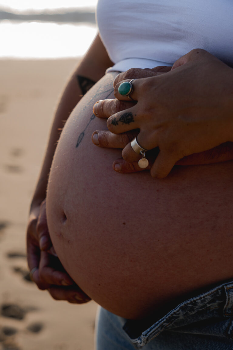 Mains d'un couple entrelacées sur un ventre de femme enceinte devant la mer sur le bassin d'arcachon pendant leur séance photo grossesse