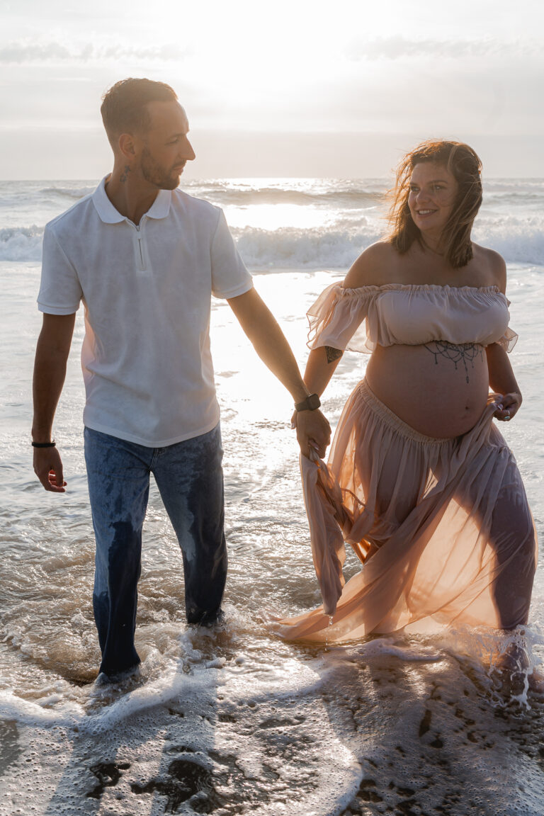Futurs parents qui se regardent en souriant, les pieds dans l'eau au coucher du soleil lors de leur séance photo grossesse à la plage de Biscarosse sur le Bassin d'Arcachon