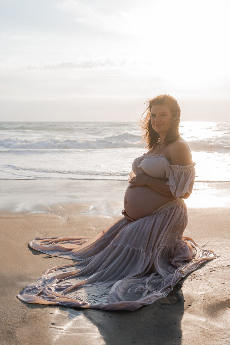 Future maman à genoux sur le sable au coucher du soleil lors de sa séance photo grossesse à la plage de Biscarosse sur le bassin d'Arcachon