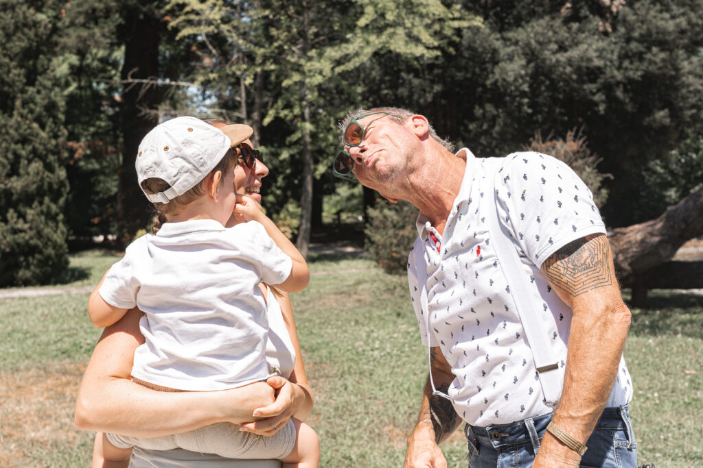 Grand-père qui fait la grimace à son petit fils dans les bras de sa maman lors de leur séance photo famille en extérieur au Parc Peixotto de Talence