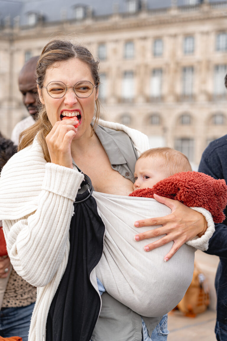 Maman qui mange un gâteau en allaitant son fils qu'elle porte en écharpe sur la place de la bourse à bordeaux