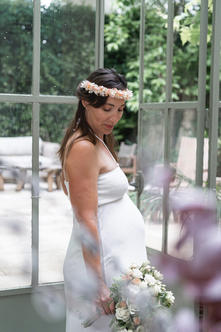 Femme enceinte qui pose dans une verrière avec une couronne de fleurs et un bouquet à la main à la maison Mandel de Bordeaux