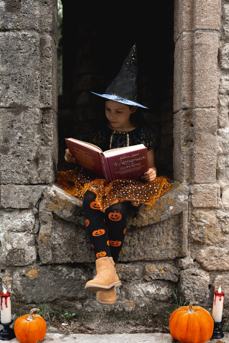 Enfant déguisée en sorcière qui lit un livre de sorcellerie assise sur un muret lors de son shooting halloween en extérieur au parc majolan de Blanquefort