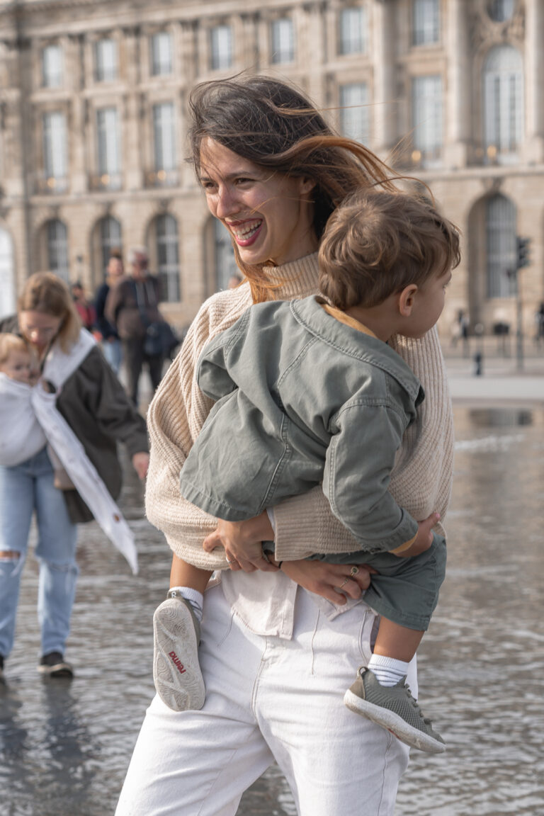 Maman qui rit avec son enfant dans les bras sur le miroir d'eau de Bordeaux pendant une séance photo en famille