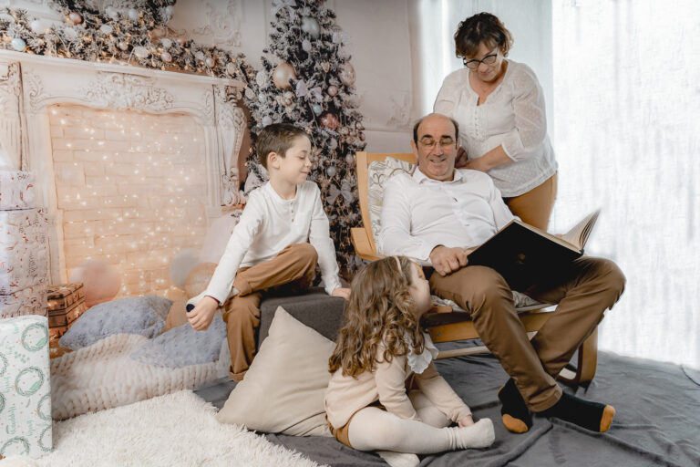 Grand-père qui lit un livre à ses petits enfants avec sa femme derrière lui lors d'un shooting photo de noël au home studio photos à Bordeaux