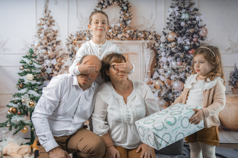 Garçon qui cache les yeux de ses grands parents pendant qu'une fille leur tend un cadeau lors de leur shooting photo noël à mon home studio photo à Bordeaux