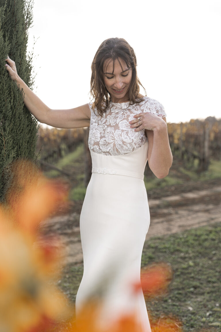 Mariée appuyée contre un arbre devant des vignes, souriant, la main posée sur le coeur lors du portrait de la mariée de sa cérémonie laïque de son mariage au chateau de courlat à Lussac