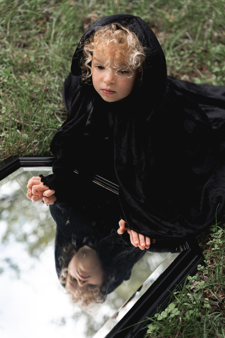 Enfant qui porte une cape qui regarde au loin accroupie devant un miroir lors d'un shooting halloween en extérieur au parc majolan de blanquefort