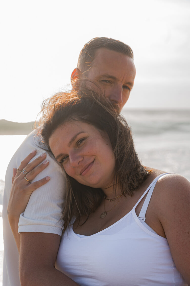 Futurs parents entrelacés qui regardent l'objectif en souriant au coucher du soleil sur la plage de biscarosse