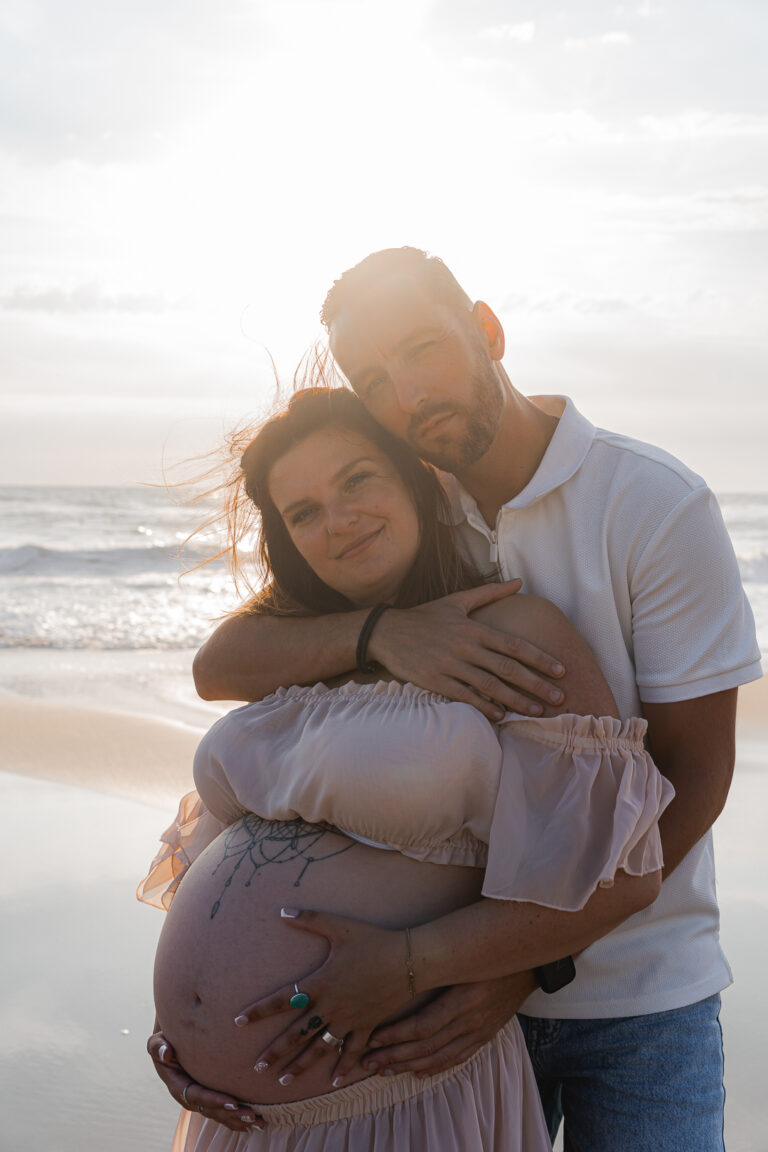 Couple enlacés qui sourit à l'objectif devant le coucher de soleil lors de leur séance photo grossesse à la plage de biscarosse